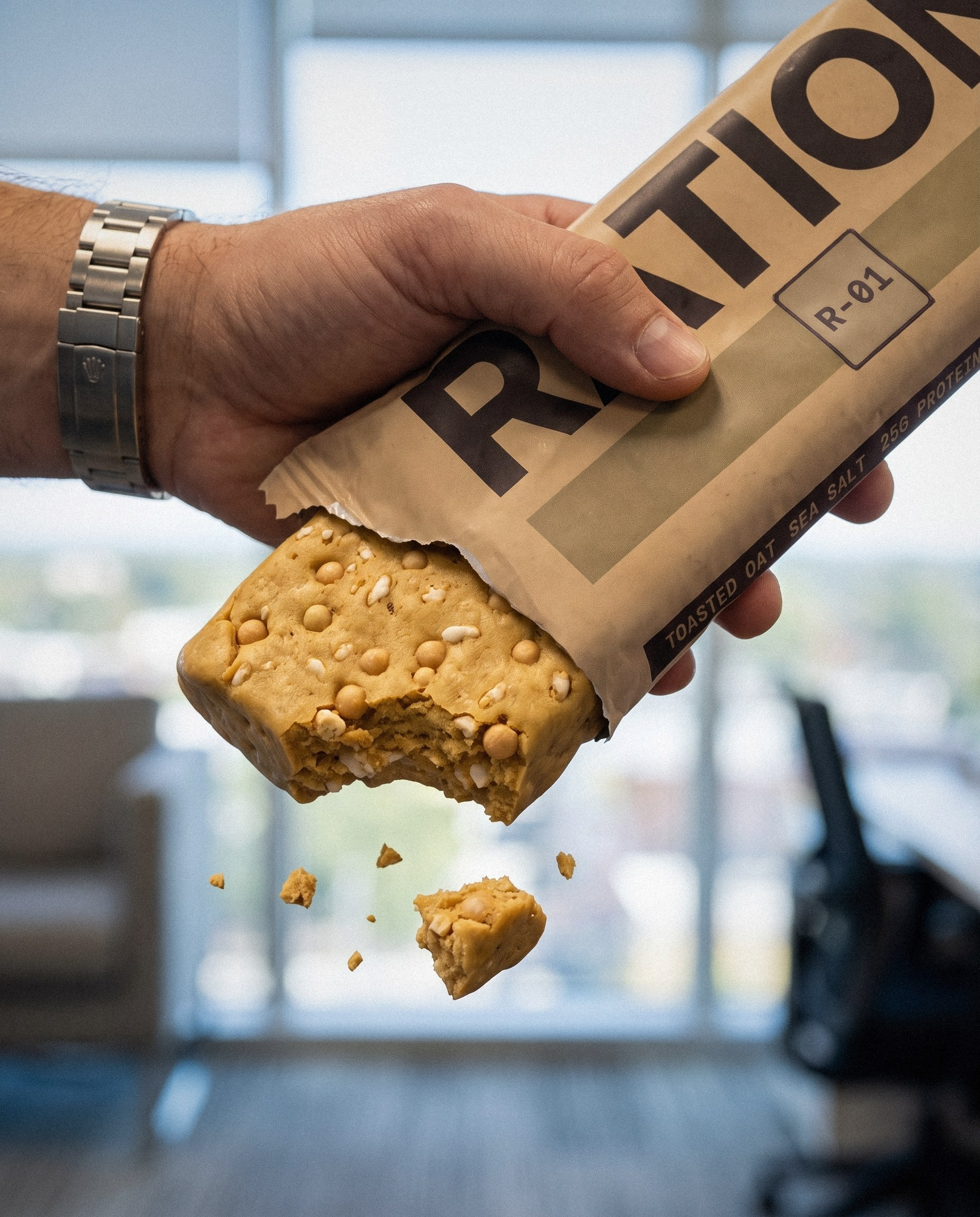 Studio render, close up: a hand holding a Ration bar with the wrapper torn halfway down, the filling, round protein crisps, and puffed rice visible at the broken edge.